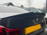 A black BMW car is parked, displaying a raised spoiler on the trunk. It's positioned in a residential driveway with trees and other vehicles nearby. The license plate is obscured.