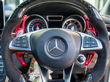 A Mercedes-AMG steering wheel with red paddle shifters, surrounded by a modern car dashboard displaying two circular gauges. The logo and buttons for audio control are visible.