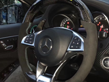 Steering wheel with carbon fiber design and buttons, displaying a Mercedes logo. Behind, illuminated speedometer gauges visible inside a car interior, with rain-covered window outside. 