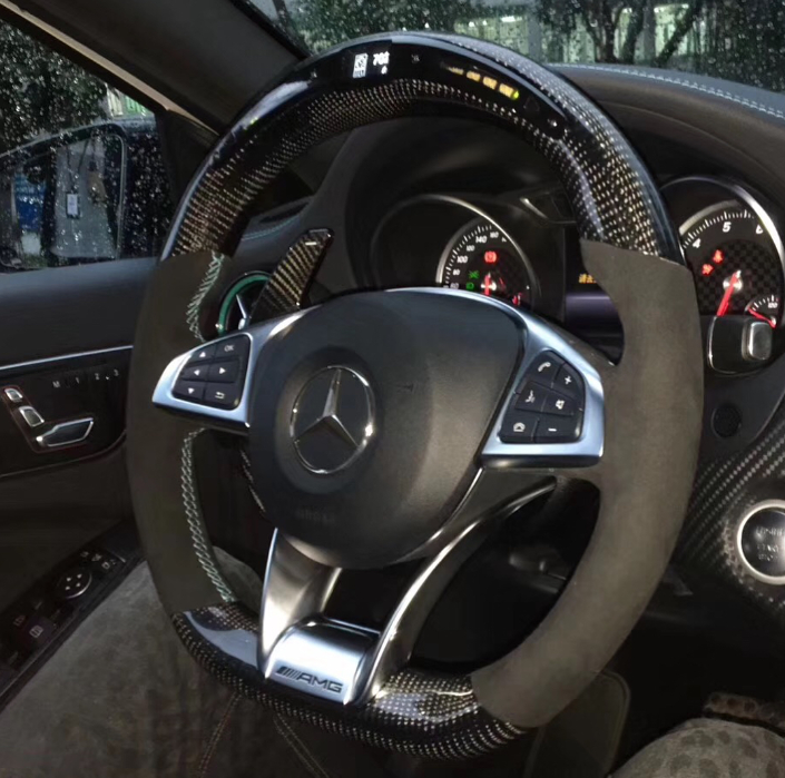 Steering wheel with carbon fiber design and buttons, displaying a Mercedes logo. Behind, illuminated speedometer gauges visible inside a car interior, with rain-covered window outside. "AMG" marked on steering wheel.