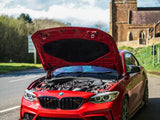 A red BMW car with its hood open reveals the engine, parked on a road near a historic stone church on a sunny day.