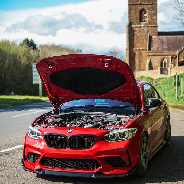 A red BMW car with its hood open reveals the engine, parked on a road near a historic stone church on a sunny day.