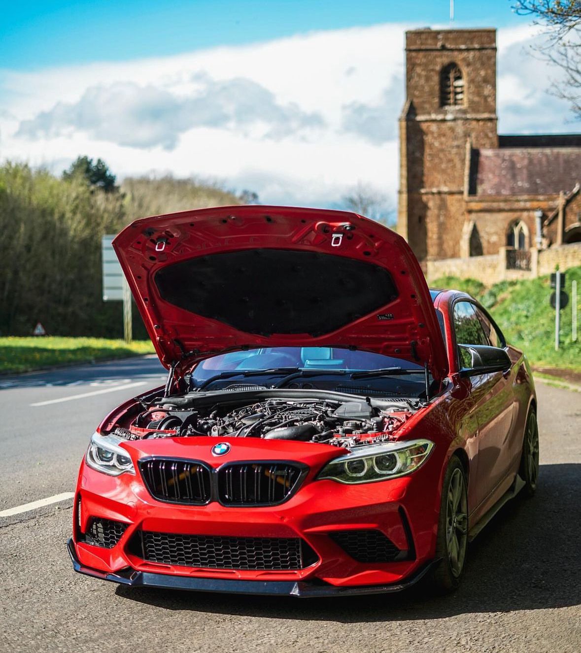 A red BMW car with its hood open reveals the engine, parked on a road near a historic stone church on a sunny day.