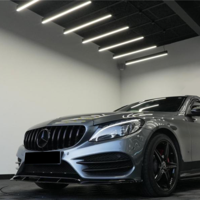 A sleek, silver car sits stationary, illuminated by bright ceiling lights in a modern showroom. The car features black alloy wheels and a prominently displayed logo on the grille.