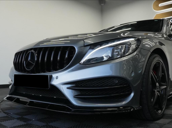 A sleek, silver Mercedes-Benz car is parked indoors on a checkered floor, showcasing its emblematic front grille, headlight design, and stylish black wheels. Bright ceiling lights reflect on the hood.