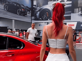 A person with red hair walks past a red car at an auto show, surrounded by large car posters and people.