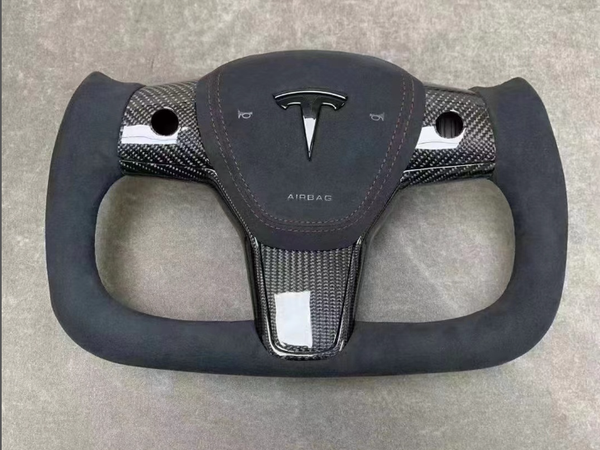 Steering yoke featuring a central emblem with two horizontal buttons. Constructed from carbon fiber and wrapped in suede, situated against a plain gray background. Text reads "AIRBAG" below the emblem.