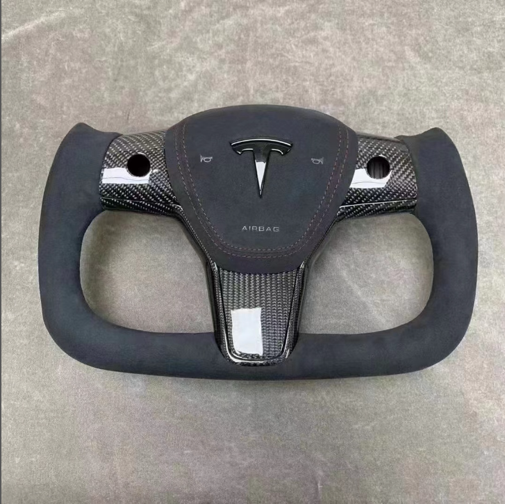 Rectangular steering wheel with a carbon-fiber design featuring the Tesla logo and "AIRBAG" text. It lies flat against a textured gray surface.