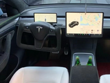 Yoke-style steering wheel with a central Tesla logo; two screens display maps and vehicle data; white seats and two green bottles in the center console; modern car interior.