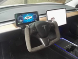 A Tesla yoke steering wheel surrounded by two digital displays in a minimalist car dashboard, featuring wood panel accents and ambient blue lighting from the center console.