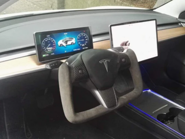 A Tesla yoke steering wheel surrounded by two digital displays in a minimalist car dashboard, featuring wood panel accents and ambient blue lighting from the center console.