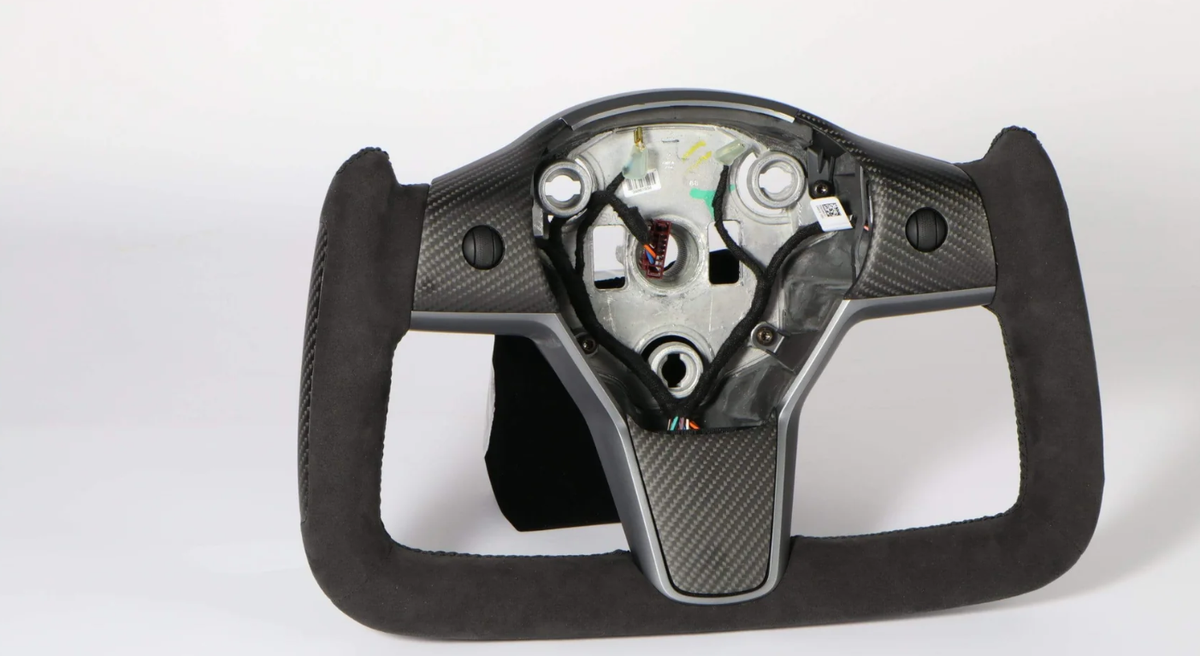 Steering wheel frame missing central cover, revealing internal mechanisms and wiring, features a carbon-fiber design and black suede grip, situated in a neutral studio setting. No text present.