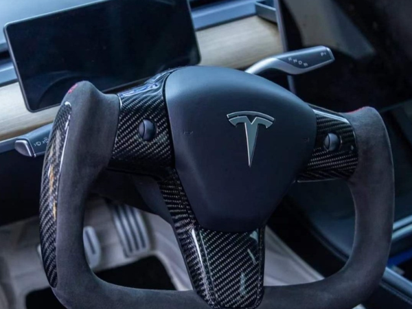 Yoke-shaped steering wheel with carbon fiber accents in a Tesla interior; dashboard display screen visible in the background, highlighting a modern, high-tech vehicle design.