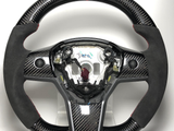 A carbon fiber steering wheel with black suede hand grips and a red top stripe is displayed on a plain white background. Center label reads 