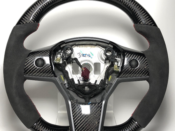 A carbon fiber steering wheel with black suede hand grips and a red top stripe is displayed on a plain white background. Center label reads "36A."