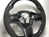 A carbon fiber steering wheel with suede grips and red stitching is displayed on a clear stand in a well-lit environment. The center is exposed, revealing internal mechanisms.