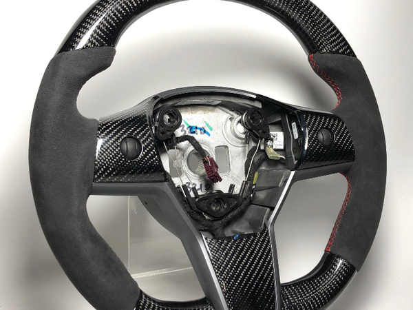 A black carbon-fiber steering wheel with suede grips and red stitching is displayed on a transparent stand, showcasing wiring and internal components in a well-lit, plain setting.