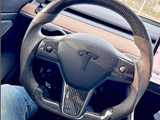 A steering wheel with a carbon fiber design displaying a Tesla logo is stationary inside the car's interior, featuring wood trim and visible driver's jeans and dashboard screen.