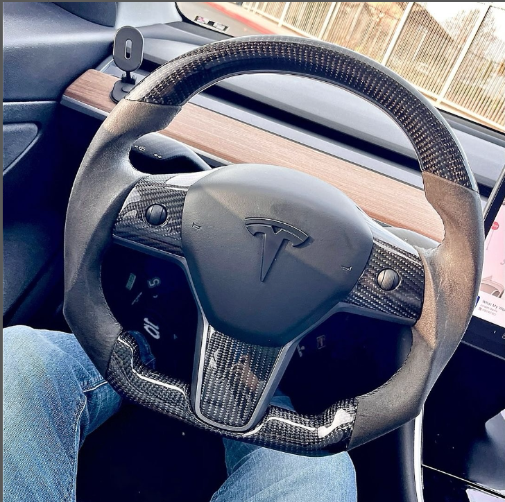 A steering wheel with a carbon fiber design displaying a Tesla logo is stationary inside the car's interior, featuring wood trim and visible driver's jeans and dashboard screen.
