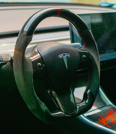 Steering wheel featuring Tesla logo, stationary in a car's cockpit. A touchscreen display is visible in the background, surrounded by a modern, sleek dashboard design.