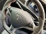 A carbon-fiber steering wheel with red stitching and control buttons, featuring a Tesla logo, is in a car interior. A digital display is visible on the dashboard behind it.