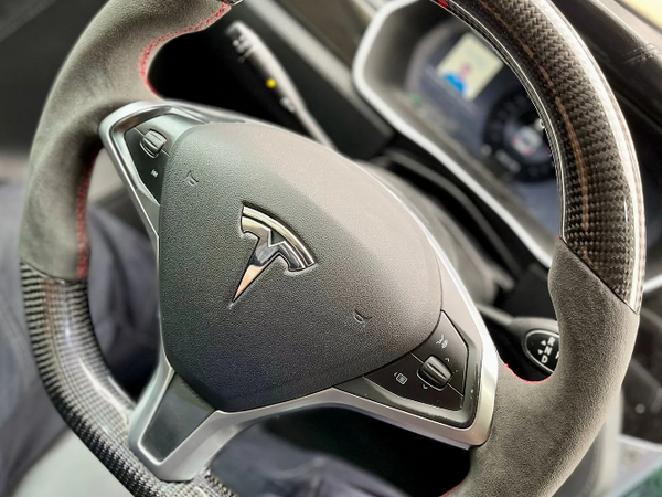 A carbon-fiber steering wheel with red stitching and control buttons, featuring a Tesla logo, is in a car interior. A digital display is visible on the dashboard behind it.