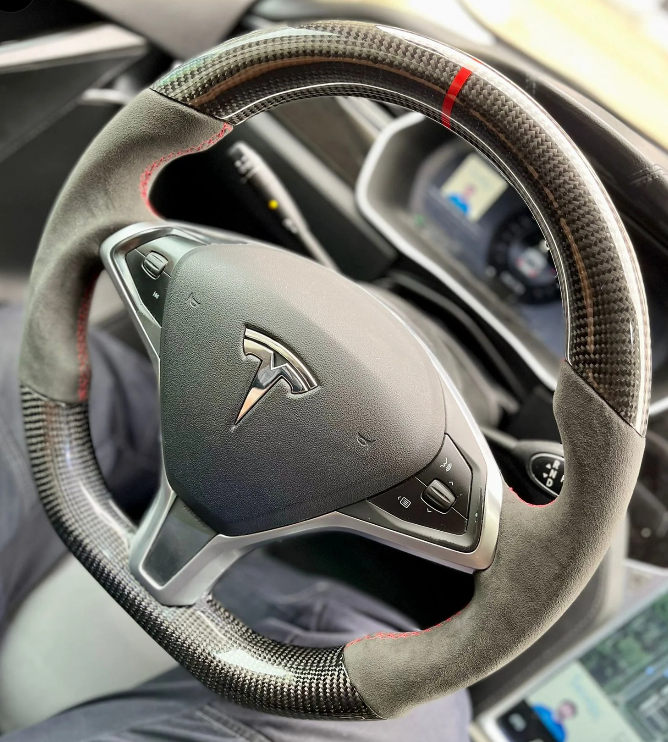 A carbon-fiber steering wheel with red stitching and control buttons, featuring a Tesla logo, is in a car interior. A digital display is visible on the dashboard behind it.