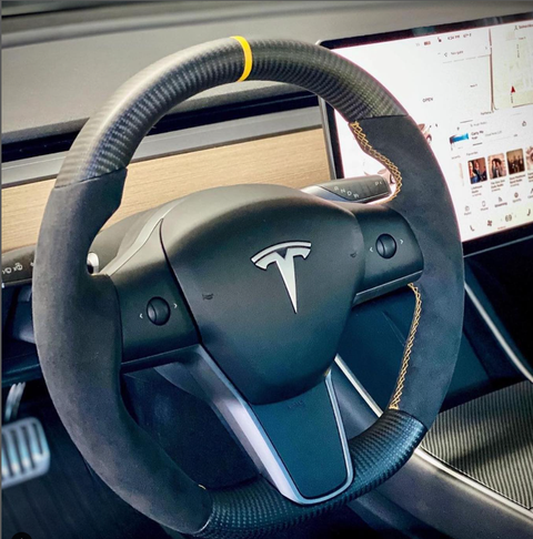 Steering wheel with yellow marker, centered in a Tesla interior featuring a large digital display screen showing a map and entertainment options, set within a modern car cabin.