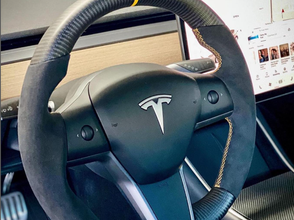 A Tesla steering wheel with a yellow top center marker is stationary inside a car, surrounded by a sleek dashboard featuring a touchscreen displaying maps and media controls.