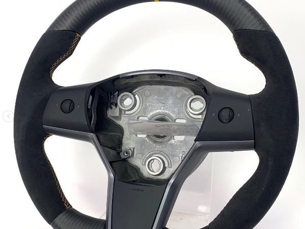 A racing steering wheel with a carbon fiber and suede grip features a yellow top centering stripe. It's positioned against a plain white background, showing exposed mounting components.
