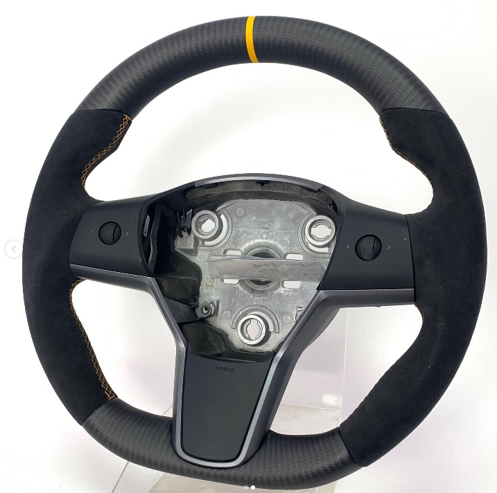 A steering wheel with a central mounting plate and integrated controls sits against a plain white background. The wheel has a black suede grip with a yellow top marker and detailed stitching.