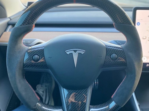 Steering wheel with carbon fiber accents and a logo centered, positioned inside a car with a wood-trimmed dashboard. A person’s legs and shoes are visible in the driver’s seat.