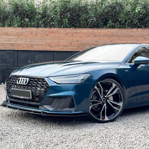 A blue car with distinctive rims is parked on a gravel surface, set against a backdrop of a wooden fence and green foliage.