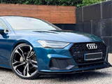 A blue Audi car is parked on a gravel surface, showcasing angular headlights and a large front grille. The background features a wooden fence and dense greenery.