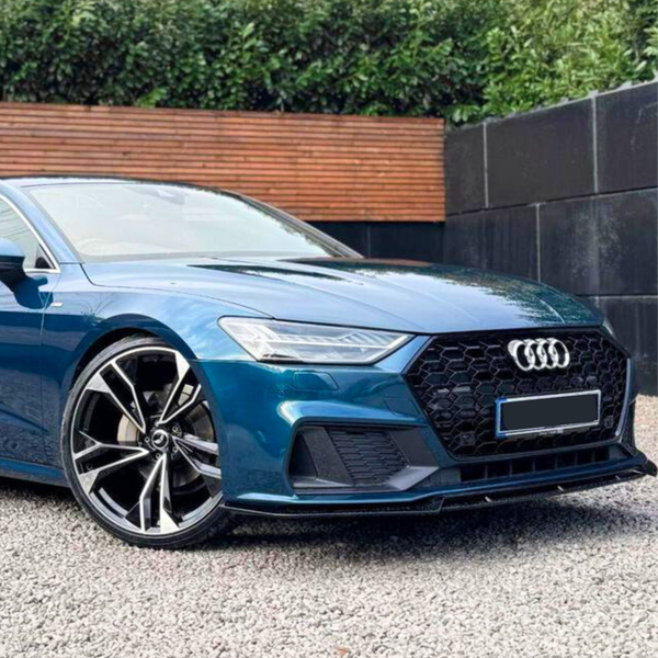 A blue Audi car is parked on a gravel surface, showcasing angular headlights and a large front grille. The background features a wooden fence and dense greenery.