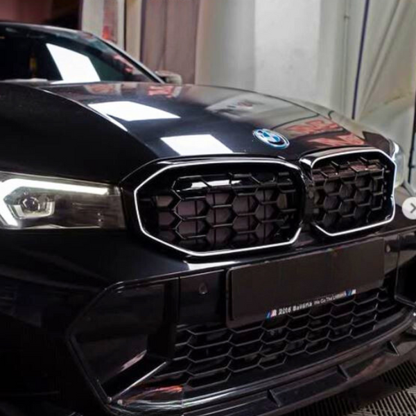 A black car's front, featuring a distinctive hexagonal grille and illuminated headlights, is parked indoors. Text on the license plate reads: "M 2Auto Bavaria. We Go The Extra Mile."
