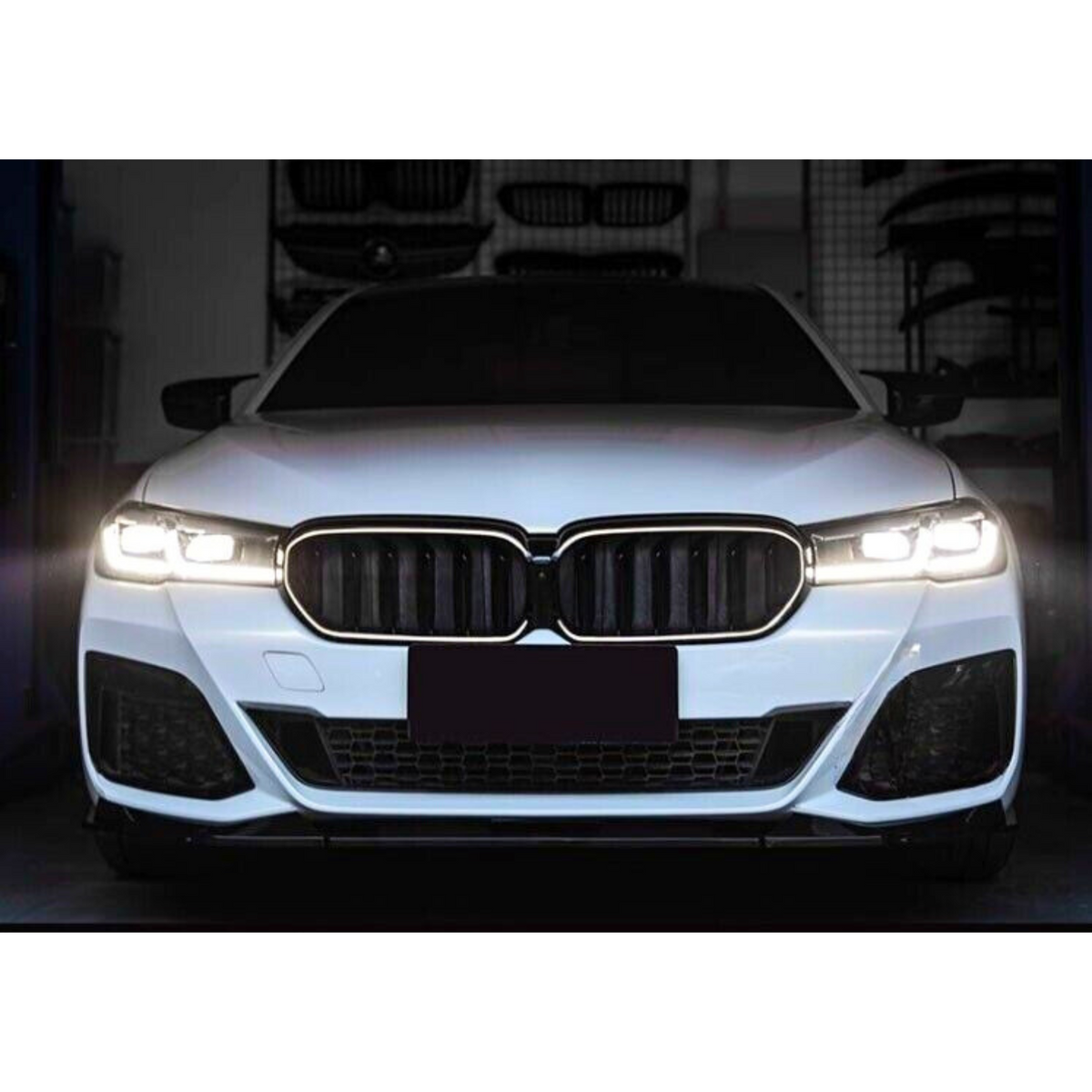 A white car with illuminated headlights is parked in a dimly lit garage, featuring a prominent grille and darkened license plate area. Various automotive parts are visible in the background.