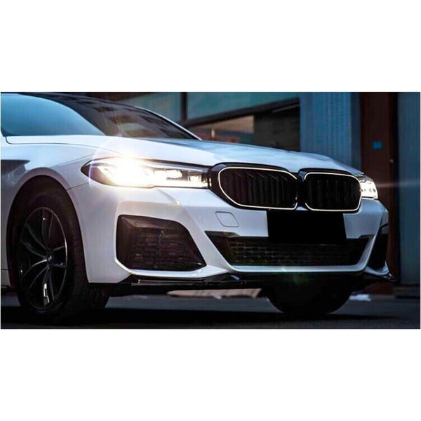A white car with illuminated headlights is parked on a street, featuring a prominent front grille and sleek design, partially surrounded by urban buildings.