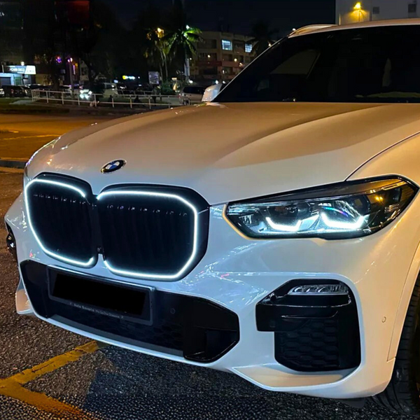 A white SUV with illuminated grille and headlights is parked on a city street at night, surrounded by blurred lights and buildings.