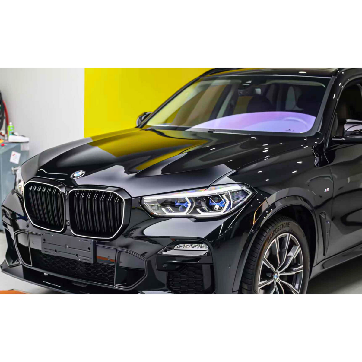 A black BMW SUV is parked indoors against a bright yellow and white wall, featuring Bridgestone tires and a sleek, modern design.
