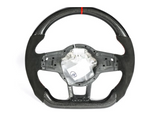 A carbon fiber and suede steering wheel with a red center stripe is displayed against a plain white background. Its center is exposed, revealing inner components, and a small plastic bag is attached.