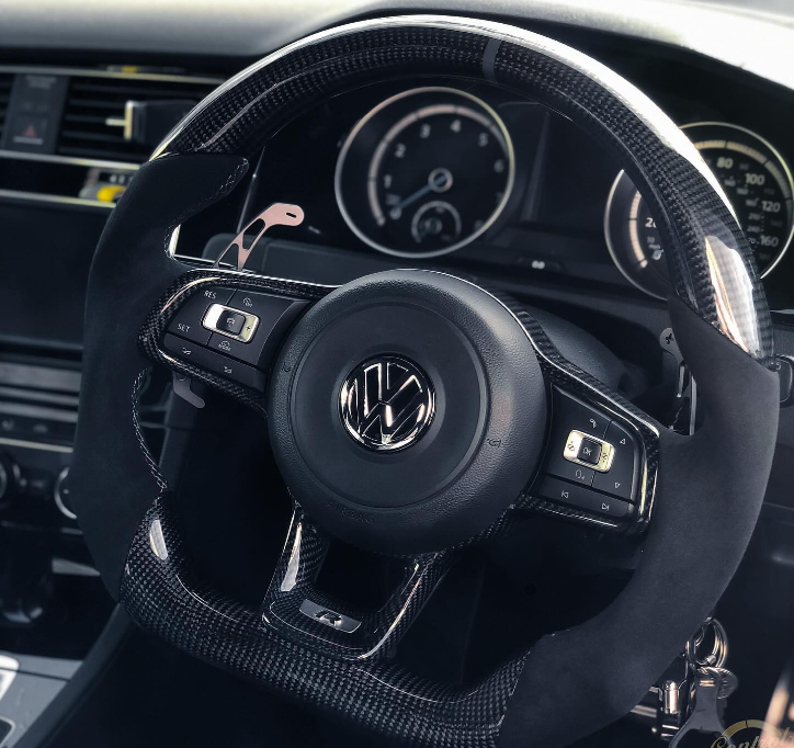 A car steering wheel with a Volkswagen logo, featuring control buttons, is set in a vehicle interior with visible speedometer and climate control dials.
