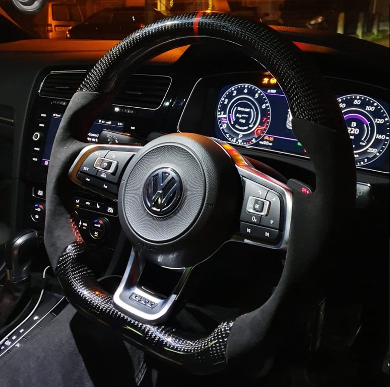 Steering wheel with Volkswagen emblem, equipped with control buttons, is positioned in front of illuminated car dashboard displaying speedometers and digital gauges in a dimly lit vehicle interior.