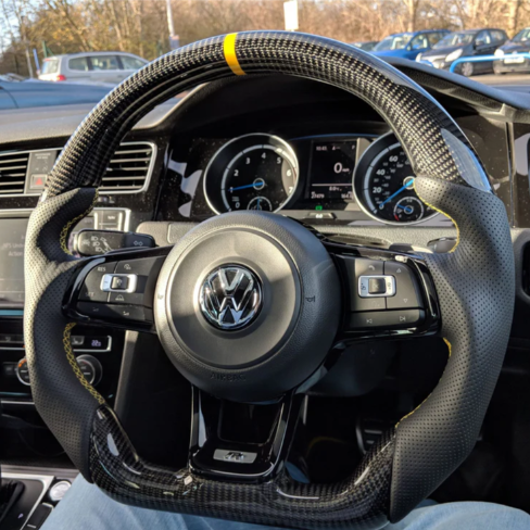 A carbon fiber steering wheel with a yellow stripe is centered in a car's interior, showing buttons and gauges. The background reveals a parking lot with several cars.