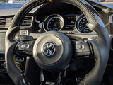 A carbon-fiber steering wheel with a yellow stripe is held by blue-jeaned legs inside a Volkswagen car. The dashboard displays various gauges. Other cars are visible through the windshield.