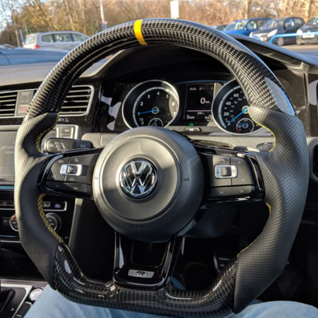 A carbon-fiber steering wheel with a yellow stripe is held by blue-jeaned legs inside a Volkswagen car. The dashboard displays various gauges. Other cars are visible through the windshield.