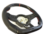 A perforated black steering wheel with red stitching and a central metallic hub is positioned on a white background, highlighting its sporty design and carbon fiber texture.