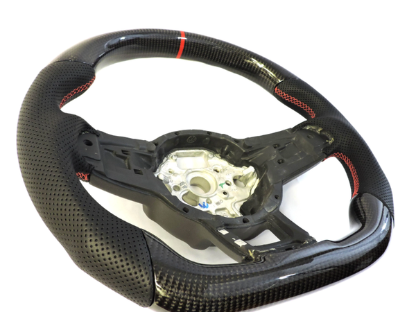 A perforated black steering wheel with red stitching and a central metallic hub is positioned on a white background, highlighting its sporty design and carbon fiber texture.