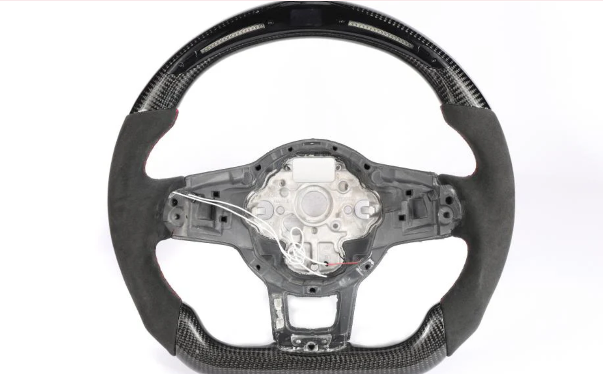 A steering wheel, partially covered in carbon fiber and black suede, has exposed wiring and a metal frame in the center, set against a plain white background.