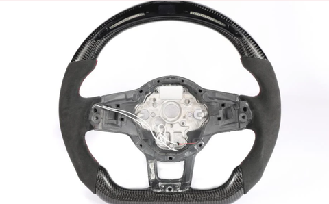 A steering wheel, partially covered in carbon fiber and black suede, has exposed wiring and a metal frame in the center, set against a plain white background.